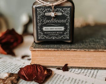 May include: A glass jar candle with a cork lid, labeled "Spellbound" with "Peach, Amber, Wild Honey" scent. The jar is tied with twine and sits on a vintage book with dried rose petals scattered around. The candle is from Midnight Muse Candles.