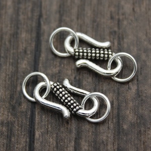 May include: Two silver-toned metal clasps with a textured design. Each clasp has a round ring on one end and a hook on the other end.