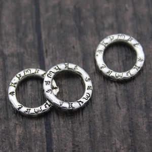 May include: Three silver-coloured metal rings with an engraved pattern of symbols around the outer edge. The rings are circular and have a slightly hammered texture. They are displayed on a dark brown wooden surface.