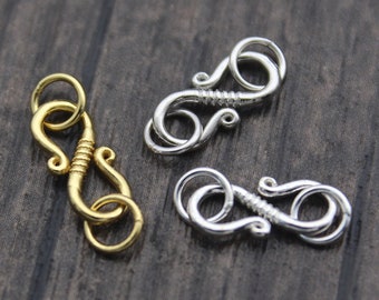 Puede incluir: Tres cierres de joyería metálicos. Uno es de color dorado y los otros dos son de color plateado. Cada cierre tiene un diseño decorativo único con cierre de gancho y ojo. Los cierres se muestran sobre una superficie de madera oscura.