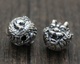 May include: Two silver-colored dragon head beads. One bead is a coiled dragon, the other is a detailed dragon head with an open mouth. The beads are on a dark wood surface.