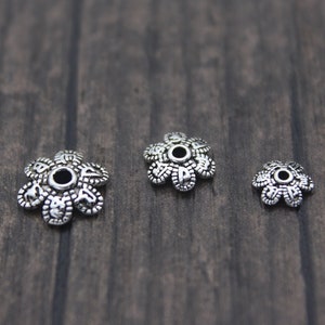 Puede incluir: Tres capuchones de cuentas plateados con forma de flor y diseños detallados de pétalos. Los capuchones varían en tamaño, con un agujero central para ensartar. Se muestran sobre una superficie oscura con vetas de madera, para bisutería.