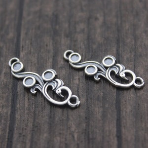 Op de afbeelding: Twee zilverkleurige metalen connector charms met een decoratief swirl-ontwerp en drie ronde accenten. De charms hebben aan elk uiteinde een lus om te bevestigen aan sieraden of andere knutselwerkjes.
