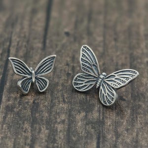 May include: Two silver butterfly-shaped ornaments. The butterflies have detailed wing patterns and are set against a dark wood grain background. The ornaments are likely jewelry or decorative items.