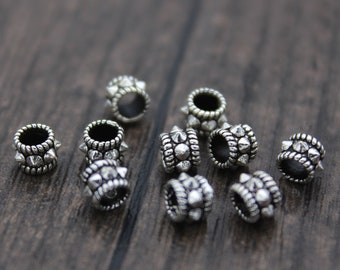 May include: Close-up of several small, silver-colored metal beads with a cylindrical shape. Each bead features a series of small, pointed spikes around the edges. The beads are scattered on a dark wooden surface.
