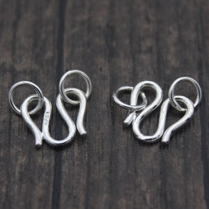 Puede incluir: Dos cierres de metal plateados con un bucle en cada extremo. Los cierres tienen forma de 'S' y están diseñados para ser utilizados como conectores para joyas.