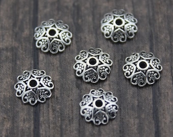 May include: Seven silver-toned metal bead caps with an intricate floral design. The caps are arranged in a semi-circle on a wood surface.