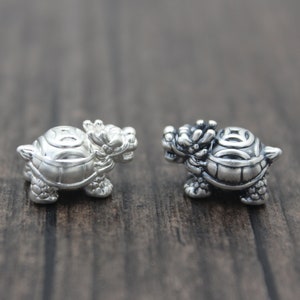 Op de afbeelding: Twee kleine, zilverkleurige schildpadfiguren met draakachtige koppen. De schildpadden hebben gedetailleerde schilden en staan op een donker houten oppervlak. De beeldjes zijn waarschijnlijk sieradenbedels of decoratieve items.
