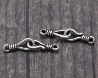 May include: Two silver-colored jewelry clasps. Each clasp has a hook and eye design, with a looped center and coiled wire detailing. The clasps are displayed on a dark wood-grain surface, showcasing their intricate design.