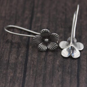 May include: A pair of silver flower earrings. Each earring has a detailed, three-dimensional flower design. The earrings are displayed on a dark brown wooden surface, highlighting their delicate craftsmanship.