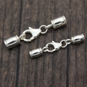 Könnte beinhalten: Zwei silberfarbene Schmuckverschlüsse mit Karabinerhaken. Jeder Verschluss hat an jedem Ende einen zylindrischen Lauf, der durch einen kleinen Ring mit dem Haken verbunden ist. Die Verschlüsse werden auf einer dunklen Holzmaserungsoberfläche gezeigt.