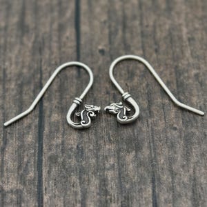 May include: A pair of silver-colored earrings with a dragon head design. The earrings have a hook style and are displayed on a wooden surface. The dragon heads are detailed with intricate patterns.