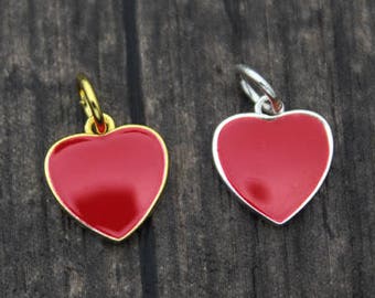 以下が含まれることがあります： 光沢のある赤い仕上げのハート型のペンダント2つ。1つは金色の縁とベイル、もう1つは銀色の縁とベイルが付いています。ペンダントは濃い木材の表面に表示されています。