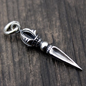 May include: A silver pendant in the shape of a stylized dagger with a decorative top. The pendant has a loop at the top for attaching to a chain.
