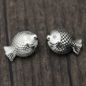 Puede incluir: Dos abalorios de pescado plateados con escamas detalladas y caras sonrientes. Los abalorios se miran.
