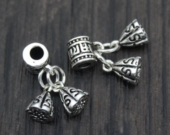 May include: Two silver-colored metal bead caps with decorative patterns. Each cap has a cylindrical component and two bell-shaped pendants connected by small chains. The beads are on a dark wood surface.