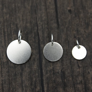 Pode incluir: Três pingentes circulares prateados de tamanhos variados. Cada pingente tem uma pequena argola na parte superior para prender a uma corrente. Os pingentes são exibidos em uma superfície de madeira escura.