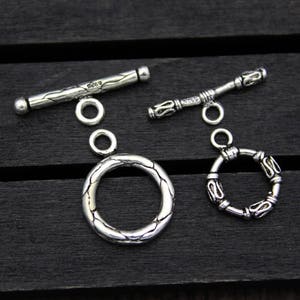 Może przedstawiać: Dwa srebrne zapięcia typu toggle do tworzenia biżuterii. Jedno zapięcie ma teksturowany pręt z napisem "Belly" i pierścień, a drugie ozdobny pręt i pierścień. Zapięcia leżą na ciemnej drewnianej powierzchni.