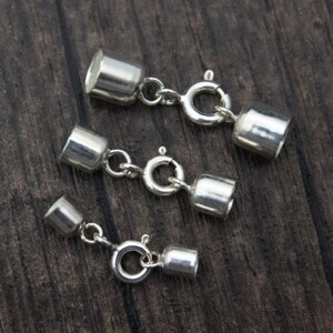 May include: Three silver-colored jewelry clasps with a cylindrical design and a spring ring clasp. The clasps are connected by small chains and are displayed on a dark wood surface.