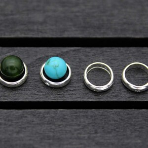 Op de afbeelding: Vier zilveren ringen met een groene en blauwe edelsteen in het midden van twee van de ringen. De ringen liggen op een donker houten oppervlak.