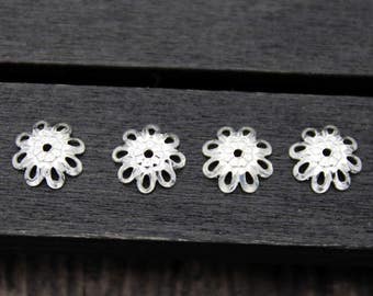 May include: Four silver-colored, flower-shaped filigree beads. Each bead has a central hole and a delicate, openwork petal design. The beads are arranged in a row on a dark wooden surface, ideal for jewelry making or craft projects.