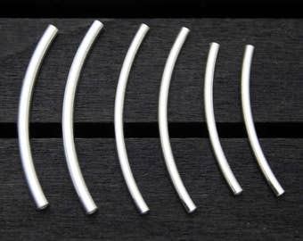 May include: Six silver-colored, curved metal bars of varying lengths are arranged on a dark wooden surface. The bars have a smooth, reflective finish and a gentle arc shape, suggesting they are jewelry components or decorative elements.