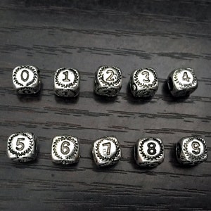 Puede incluir: Cuentas de metal plateado, cada una con un número diferente del 0 al 9. Los números están en una fuente clásica y rodeados por un borde decorativo. Las cuentas son cuadradas y tienen un agujero para ensartar.