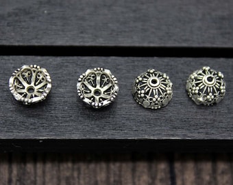 May include: Four silver-colored metal bead caps with intricate openwork designs. The caps are arranged in a row on a dark wooden surface. Each cap has a unique pattern, showcasing detailed craftsmanship. These decorative bead caps are ideal for jewelry making.