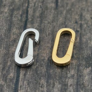 May include: Two metal clasps, one silver and one gold, are displayed against a dark wood grain background. The silver clasp is a carabiner style, while the gold one is an oval shape. These clasps are likely used for jewelry or crafting.