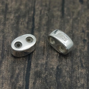 May include: Two small, oval-shaped silver beads with two holes each, lying on a dark wooden surface. The beads have a shiny finish and are marked with "925". These beads are suitable for jewelry making.