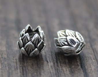 Puede incluir: Dos cuentas de metal plateadas con forma de alcachofas. Las cuentas están detalladas con hojas texturizadas y se muestran sobre una superficie de madera.