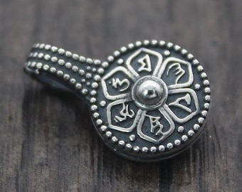 May include: A silver pendant with a circular design featuring a central sphere and six stylized petals, surrounded by a dotted border. The pendant has a small, curved handle. The background is a dark wood surface.