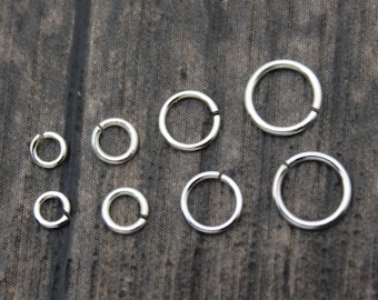 May include: A set of nine silver jump rings of varying sizes, arranged on a dark wood surface.