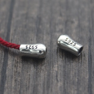 Puede incluir: Primer plano de un cordón rojo con dos cierres plateados. Cada cierre está grabado con "S925". Los cierres están separados, mostrando el cordón. El fondo es una superficie de madera oscura.