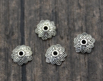 May include: Four silver metal flower-shaped bead caps with a hole in the center. The bead caps are on a wooden surface.