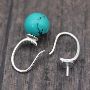 May include: Two silver-colored earring components. One features a turquoise-colored, round bead. The other component is a simple hook with a post. The earrings are displayed on a dark wood surface.