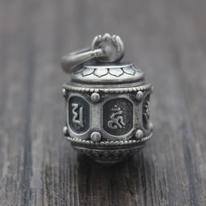 Puede incluir: Péndulo de plata en forma de rueda de oración con intrincados detalles y escritura tibetana. La rueda está diseñada para girar y es un símbolo popular de buena suerte y fortuna.