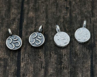 May include: Four small, circular silver-toned metal charms, each featuring an embossed Om symbol on the front surface. Every charm includes a small loop at the top for attachment, displaying a weathered, oxidized finish with dark recessed details.