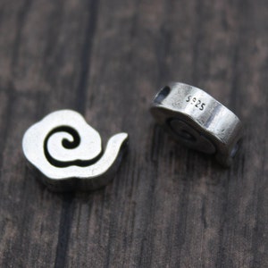 Puede incluir: Dos cuentas de plata con forma de nube con un remolino en su interior. La cuenta de la derecha tiene la inscripción "S925" en el lateral.