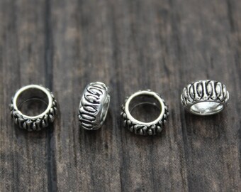 May include: Four silver-colored, round beads with a detailed design. Each bead has a ring shape with a textured surface and a pattern of vertical lines and small, rounded shapes. The beads are displayed on a dark wooden surface.