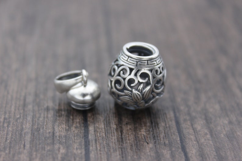 Puede incluir: Un charm de plata en forma de peque&ntilde;o frasco redondo adornado con tapa. El frasco tiene un dise&ntilde;o floral y est&aacute; abierto en la parte superior. La tapa est&aacute; separada y tiene un bucle para sujetarlo a una cadena.