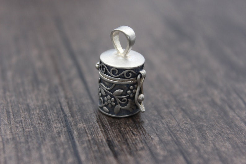 May include: A silver charm in the shape of a small, ornate box with a hinged lid. The box is decorated with a floral design and has a loop at the top for attaching to a necklace or bracelet.
