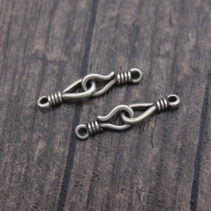 Op de afbeelding: Twee zilverkleurige metalen sluitingen met een haak- en oogontwerp. Elke sluiting heeft een lusvormig uiteinde en een haak die verbinding maakt met een lus. De sluitingen liggen op een donker oppervlak met houtnerf.