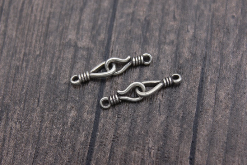 Op de afbeelding: Twee zilverkleurige metalen sluitingen met een haak- en oogontwerp. Elke sluiting heeft een lusvormig uiteinde en een haak die verbinding maakt met een lus. De sluitingen liggen op een donker oppervlak met houtnerf.