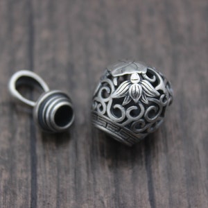 Puede incluir: Pendiente de plata con un dise&ntilde;o redondo calado que presenta una flor de loto y espirales intrincados. El pendiente tiene una tapa separada con un anillo para sujetarlo a una cadena.