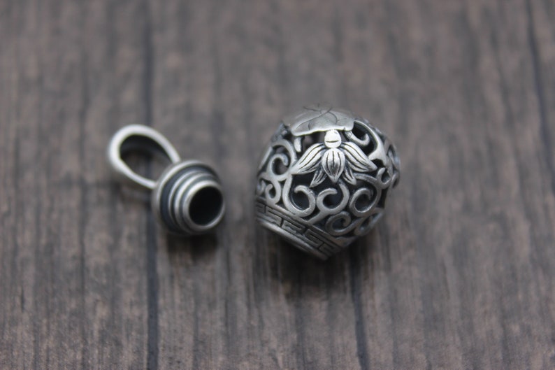 Puede incluir: Pendiente de plata con un dise&ntilde;o redondo calado que presenta una flor de loto y espirales intrincados. El pendiente tiene una tapa separada con un anillo para sujetarlo a una cadena.