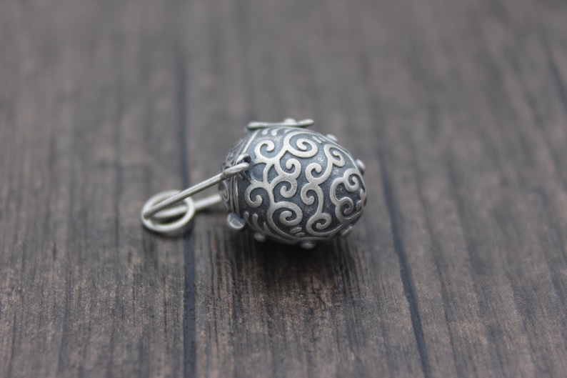 May include: A silver charm in the shape of a small, ornate egg with a swirl design. The charm has a small loop at the top for attaching to a necklace or bracelet.