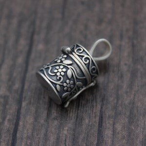 May include: A silver charm in the shape of a small, ornate box with a floral design. The box has a hinged lid and a loop for attaching to a necklace or bracelet.