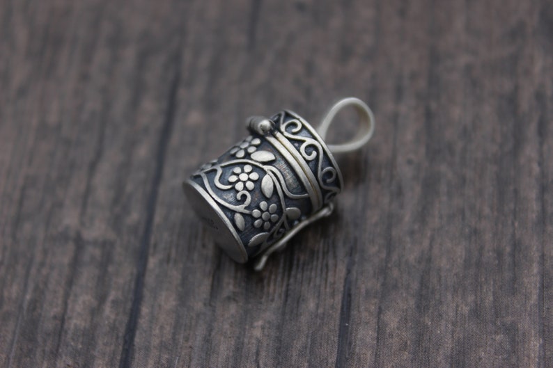 May include: A silver charm in the shape of a small, ornate box with a floral design. The box has a hinged lid and a loop for attaching to a necklace or bracelet.