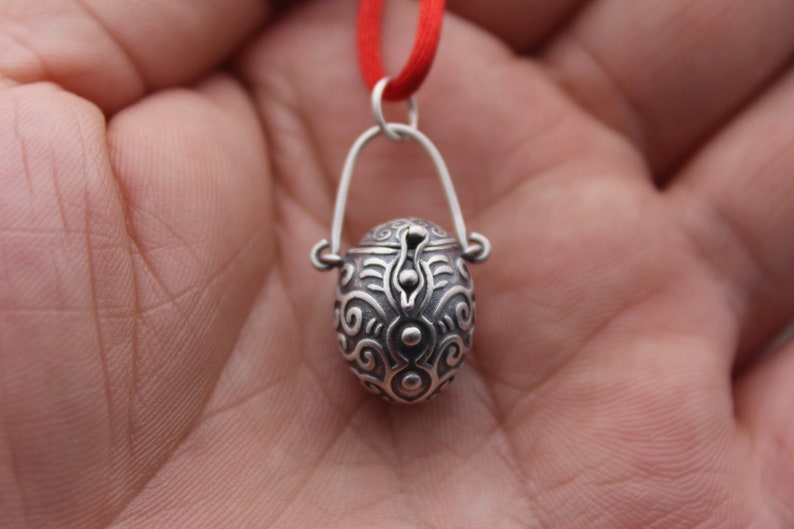 May include: A silver pendant necklace with a small, ornate, hinged box charm. The box is decorated with a swirling, abstract pattern.